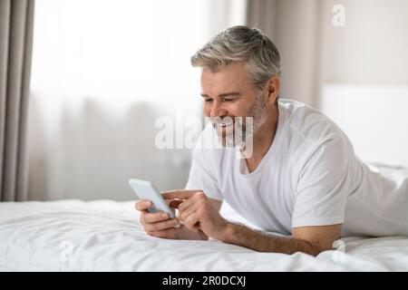 Positiver erwachsener Mann, der zu Hause im Bett ruht, telefoniert Stockfoto