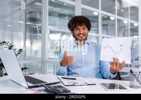 Portrait von jungen erfolgreichen Finanzierern, die im Büro arbeiten, lächelnd und vor die Kamera schauen, zeigt Grafik mit positiver Dynamik der wirtschaftlichen Gewinne, Geschäftsmann bei der Arbeit mit einem Notebook. Stockfoto