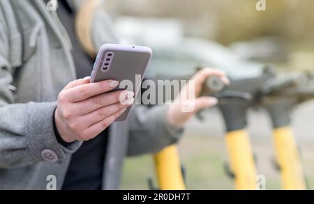 Die junge Frau entsperrt einen E-Roller mit ihrem Handy, dem modernen Transport in der Großstadt Stockfoto