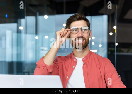 Porträt eines erfolgreichen und lächelnden jungen Mannes mit Bart und rotem Hemd, der im Büro sitzt, an einem Laptop arbeitet, selbstbewusst zur Seite schaut und seine Brille mit der Hand hält. Stockfoto