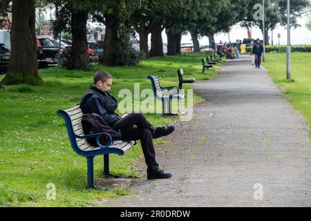Ein junger Mann, der allein auf einer Parkbank sitzt und Musik über seine Kopfhörer hört Stockfoto