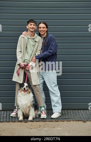 Fröhlicher schwuler Mann mit Zöpfen, die lächelnden tätowierten Freund umarmen, in lässigem Outfit, mit Leine eines australischen Schäferhundes, der nebenan stand Stockfoto