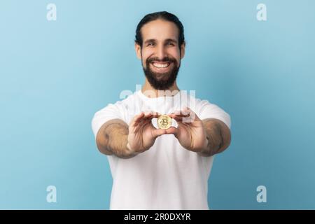 Porträt eines zufriedenen Mannes mit Bart, der ein weißes T-Shirt trägt und Bitcoin hält, und auf die neue digitale Kryptowährung achtet. Innenstudio, isoliert auf blauem Hintergrund. Stockfoto