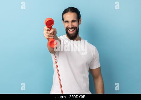 Porträt eines gutaussehenden, glücklichen Mannes mit Bart, der weißes T-Shirt trägt und den Retro-Telefonhörer vor der Kamera zeigt, und fragt, ob er ans Telefon gehen will. Innenstudio, isoliert auf blauem Hintergrund. Stockfoto