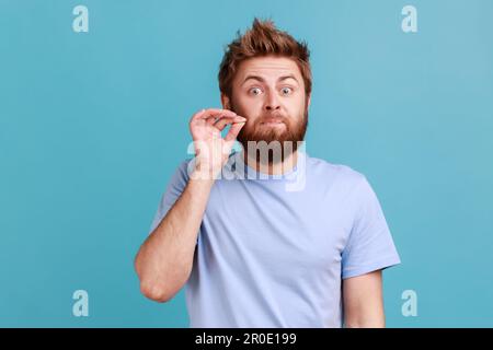 Ich erzähle es niemandem. Porträt eines gutaussehenden jungen, bärtigen Mannes, der den Mund aufmacht, geheime Informationen bewahrt, in die Kamera schaut. Innenstudio, isoliert auf blauem Hintergrund. Stockfoto