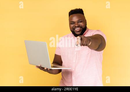 Porträt eines optimistischen bärtigen Mannes, der ein rosa Hemd trägt, einen Laptop hält, mit dem Finger auf die Kamera zeigt und lächelt, dich auswählt, freiberufliche Stellenanzeige. Innenstudio-Aufnahme isoliert auf gelbem Hintergrund Stockfoto