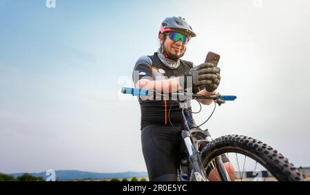 Männlicher Radfahrer mit Helm und Handy. Radfahrer auf seinem Fahrrad, der im Freien SMS mit dem Telefon schreibt. Radfahrer mit Helm und Brille, der am Handy SMS schreibt Stockfoto