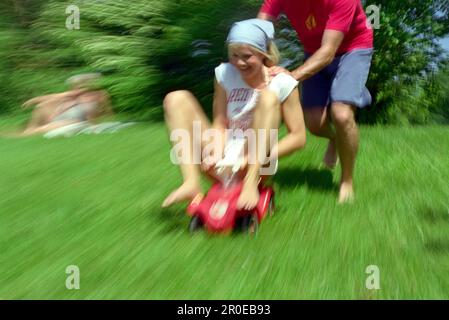 Ein junger Mann schubst eine junge Frau auf ein Spielzeugauto Stockfoto