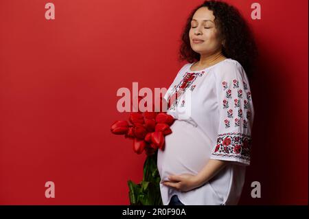 Entzückende Erwachsene schwangere Frau in ukrainischer ethnischer Kleidung, hält Bauch, posiert mit roten Tulpen, isolierter Hintergrund Stockfoto