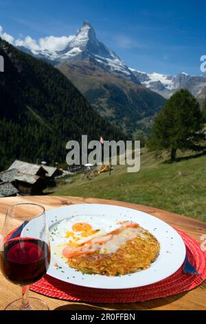 Rösti mit Spiegeleiern auf einem Teller und einem Glas Rotwein serviert in einem Bergrestaurant, Findeln, Matterhorn (4478 m) im Hintergrund, Zermatt, Wallis, Stockfoto