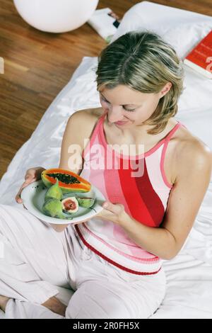 Junge Frau, die sich im Bett mit einem Teller mit exotischen Früchten entspannt Stockfoto