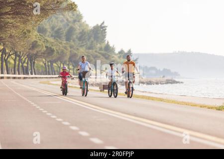 Zwei Kinder mit Helmen auf dem Kopf und lächelnde Eltern, die auf einer familienfreundlichen Radroute entlang der Küste mit Blick auf die Küste Fahrräder fahren. Stockfoto