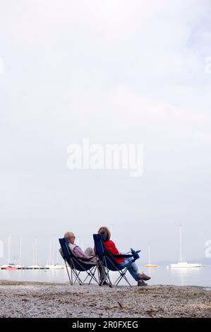 Älteres Ehepaar, das auf Klappstühlen am Strand am Ammersee in Bayern sitzt Stockfoto