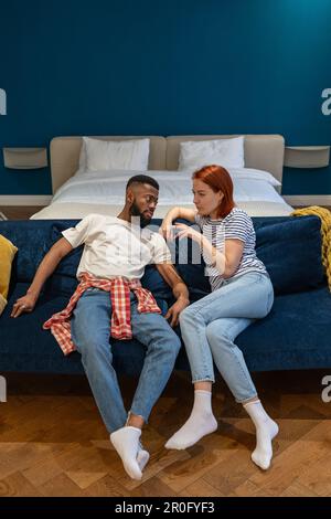 Konkurrierendes, multiethnisches gestresstes Paar, das auf der Couch sitzt und über Probleme spricht. Stockfoto