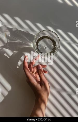 Weibliche Hand berührt sanft ein Glas klares Wasser, Blick von oben, abstrakte Schatten, Morgenlicht Stockfoto
