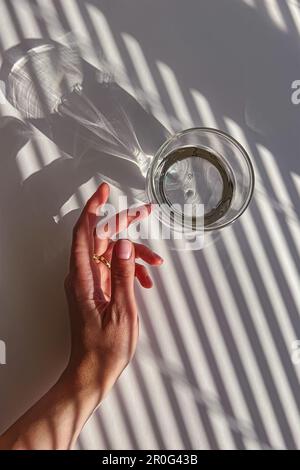 Weibliche Hand berührt sanft ein Glas klares Wasser, Blick von oben, abstrakte Schatten, Morgenlicht Stockfoto