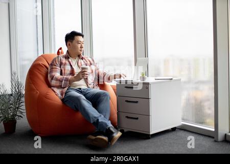 Ein fleißiger asiatischer Angestellter in blauen Hosen und kariertem Hemd, der eine Tasse köstlichen Kaffees hält und drinnen auf dem Laptop tippt. Junger IT-Mann, der Websites mit moderner Drahtlostechnologie programmiert. Stockfoto