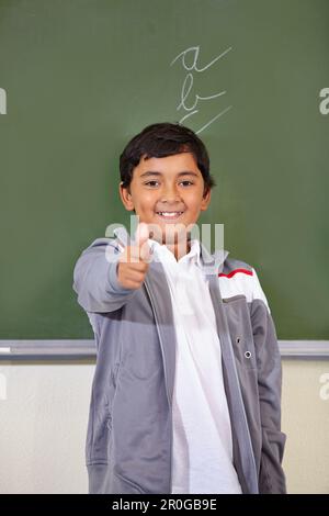 Porträt, Kind und Daumen hoch von einem Schüler auf einer Tafel im Klassenzimmer, in der Grundschule und im Modell. Tafel, Bildung und Kind mit Handgeste für Stockfoto