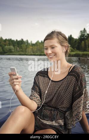 Junge Frau, die Musik hört, während sie auf einem Steg am Starnberger See in Bayern sitzt Stockfoto