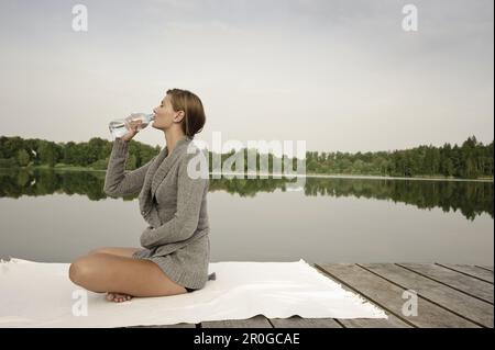 Junge Frau, die eine Flasche Wasser trinkt, während sie auf einem Steg am Starnberger See in Bayern sitzt Stockfoto