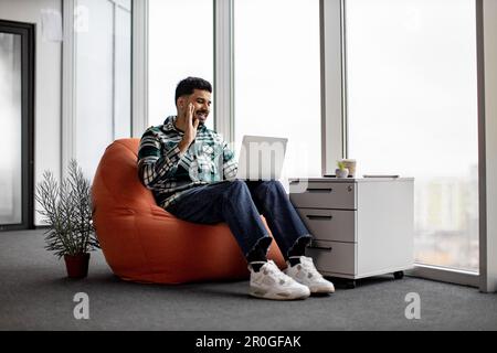 Ein gutaussehender, fröhlicher hindu-Mann, der Videokonferenzen über ein drahtloses Gerät führt und Kollegen die Hand winkt. Ein Mann mit Bart in Jeans, Hemd und weißen Sneakers teilt seine Erfahrung mit Kollegen im Büro. Stockfoto