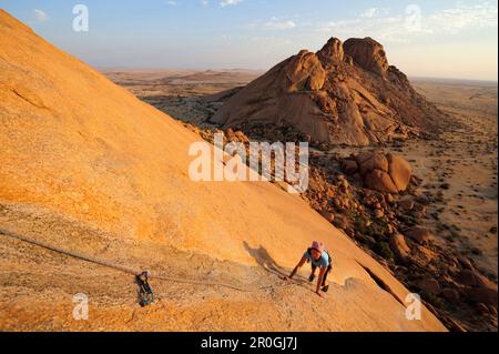 Frau Klettern am roten Fels, Sugarloaf im Hintergrund, Große Spitzkoppe, Namibia Stockfoto