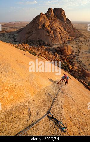 Frau Klettern am roten Fels, Sugarloaf im Hintergrund, Große Spitzkoppe, Namibia Stockfoto