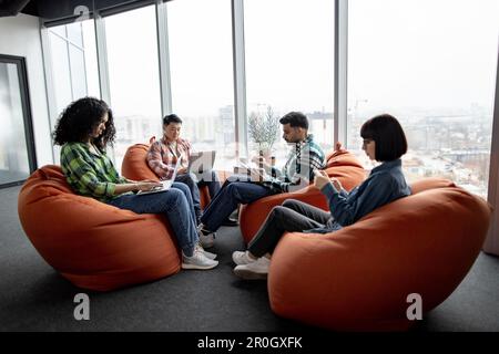 Gruppe junger Geschäftsleute in lässiger Kleidung mit modernen Geräten, während sie auf Terrakotta-Sitzsäcken im Gemeinschaftsarbeitsbereich sitzen. Multikulturelle Teamkollegen, die nach Informationen zu einem neuen Projekt suchen. Stockfoto