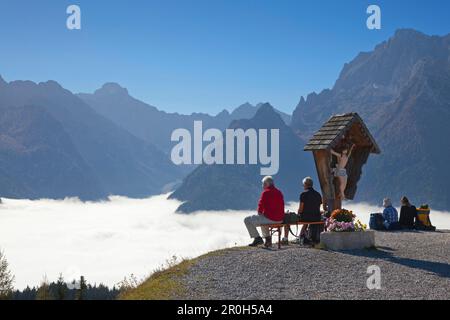 Wanderer, die sich an einem Wegkreuz ausruhen, Blick über den Nebel im Tal auf Hochkalter, Berchtesgaden Region, Berchtesgaden Nationalpark, Oberbayern, Stockfoto