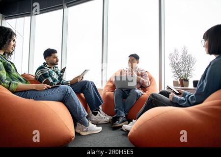 Selbstbewusster asiatischer Mann in Freizeitkleidung, der mit multiethnischen Kollegen spricht, während er in einem modernen Büro mit einem Computer sitzt. Professionelle Führungskraft, die ein intensives Brainstorming am kreativen Arbeitsplatz durchführt. Stockfoto