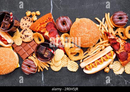 Tischszene mit Junk-Food, verstreut über einen dunklen Hintergrund. Mischung aus Essen zum Mitnehmen und Fast Food. Pizza, Hamburger, pommes frites, Pommes frites, Hot Dogs, Süßigkeiten. Stockfoto