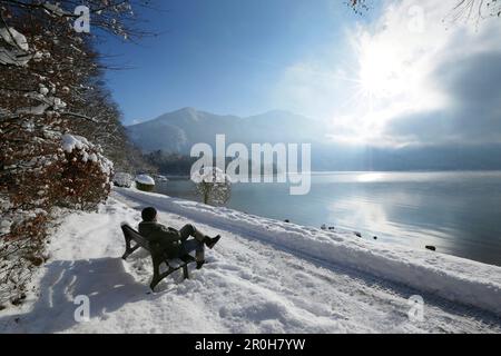 Man sitzt auf einer Bank am verschneiten See promenade, See Kochel, Kochel, Oberbayern, Deutschland Stockfoto