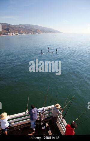 USA, Kalifornien, Malibu, Männer fischen am Malibu Pier, während vier Paddleboarder auf ruhiger See vorbeifahren Stockfoto