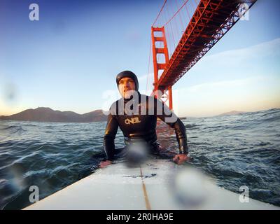 USA, Kalifornien, San Francisco, Fort Point, Surfer auf seinem Surfbrett unter der Golden Gate Bridge bei Sonnenuntergang Stockfoto