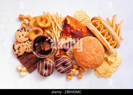 Tischszene mit Junk-Food. Kollektion auf weißem Marmorhintergrund verstreut. Mischung aus Essen zum Mitnehmen und Fast Food. Pizza, Hamburger, pommes Frites, Pommes Frites Stockfoto