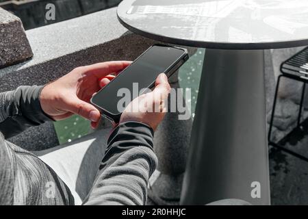 Ein Mann mit einem Smartphone in der Hand sitzt mitten in der Stadt bei Tageslicht Stockfoto
