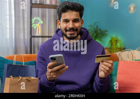 Glücklicher, einkaufsamer, indischer Konsumenten, der mit Einkaufstaschen in einem modernen Apartment sitzt und online bestellt, mit Kreditkarte bezahlt. Rabattangebot im Internet. Junger hindu-Typ auf dem Sofa Stockfoto