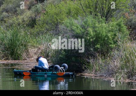 Ein Mann, der in einem kleinen Boot am Ufer angeln will, umgeben von hohen Bäumen und üppigen Flusspflanzen, die an einem sonnigen Tag wachsen. Stockfoto