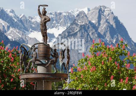 Brunnen mit Skulpturen des traditionellen Coopers Dance, Wetterstein Mountains, Zugspitze, Kastanienblüte im Frühling, Partenkirchen, Garmisch-Partenkirc Stockfoto