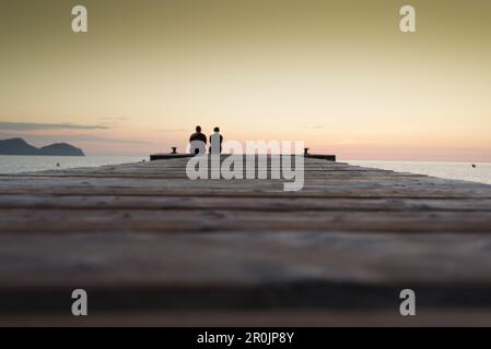 Ein Paar, das am Ende eines langen Piers in Morgenstimmung sitzt. Playa de Muro Beach, Alcudia, Mallorca, Balearen, Spanien Stockfoto