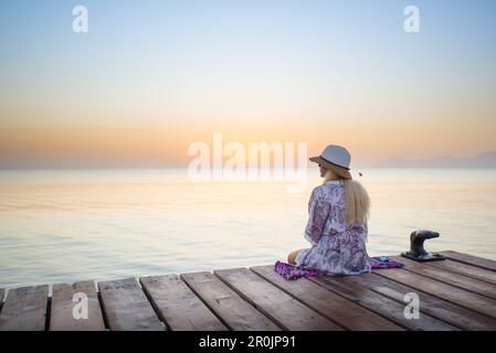 Junge blonde Frau, die am Ende eines langen Piers in Morgenstimmung sitzt und den Blick auf den Sonnenaufgang genießt. Playa de Muro Beach, Alcudia, Mallor Stockfoto