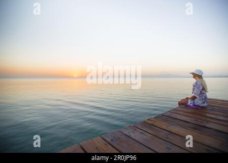 Junge blonde Frau, die am Ende eines langen Piers in Morgenstimmung sitzt und den Blick auf den Sonnenaufgang genießt. Playa de Muro Beach, Alcudia, Mallor Stockfoto