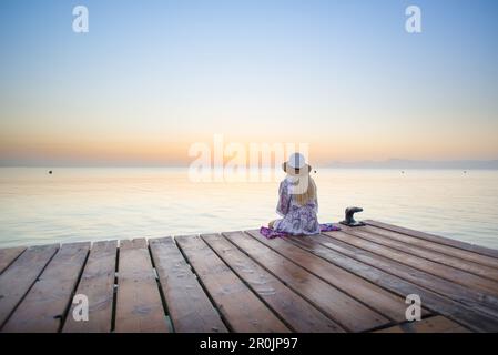 Junge blonde Frau, die am Ende eines langen Piers in Morgenstimmung sitzt und den Blick auf den Sonnenaufgang genießt. Playa de Muro Beach, Alcudia, Mallor Stockfoto