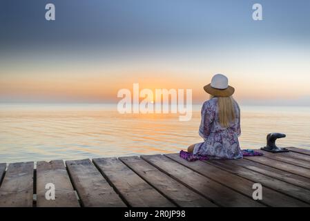 Junge blonde Frau, die am Ende eines langen Piers in Morgenstimmung sitzt und den Blick auf den Sonnenaufgang genießt. Playa de Muro Beach, Alcudia, Mallor Stockfoto