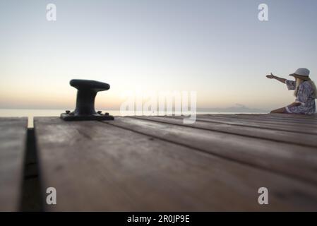 Junge Frau, die am Ende eines langen Piers in Morgenstimmung sitzt und auf das Mittelmeer zeigt. Playa de Muro Beach, Alcudia, Mallorca, Bal Stockfoto