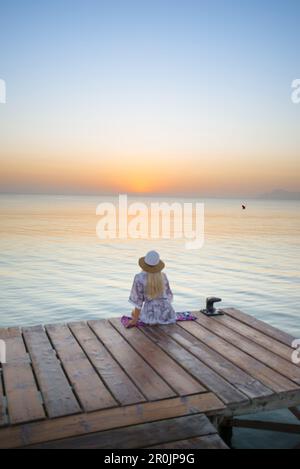 Junge blonde Frau, die am Ende eines langen Piers in Morgenstimmung sitzt und den Blick auf den Sonnenaufgang genießt. Playa de Muro Beach, Alcudia, Mallor Stockfoto