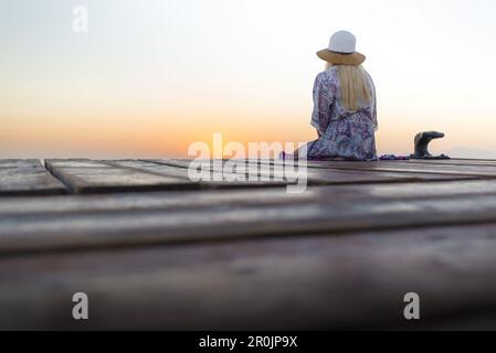 Junge blonde Frau, die am Ende eines langen Piers in Morgenstimmung sitzt und den Blick auf den Sonnenaufgang genießt. Playa de Muro Beach, Alcudia, Mallor Stockfoto