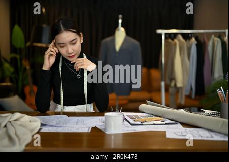 Fokussierte asiatische Modedesignerin oder Schneiderin, die in einem Studio an ihrer neuen Bekleidungskollektion arbeitet. Stockfoto
