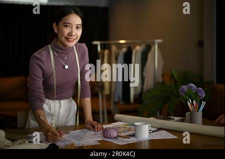 Lächelnde und charmante Millenials asiatische Modedesignerin ist in ihrem Studio, steht an ihrem Tisch. Stockfoto