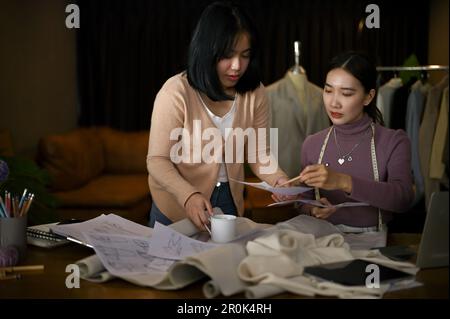 Fokussierte und professionelle asiatische Modedesignerin plant ihre neue Kollektion mit ihrer Assistentin in einem Kreativstudio. Stockfoto